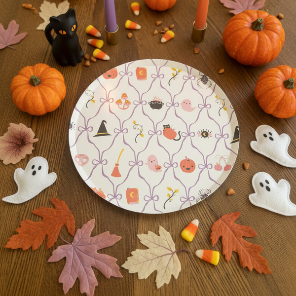 A hand holding a round white paper plate with a purple lattice pattern and Halloween-themed illustrations, set against a wooden tabletop.