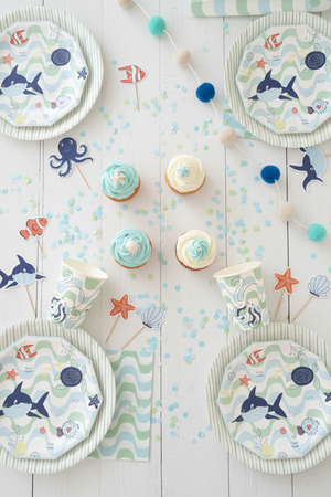 Underwater-themed party tableware set including a paper plate, matching napkins, paper cups, pom-pom garland, and fish-shaped toppers on a wooden tabletop.