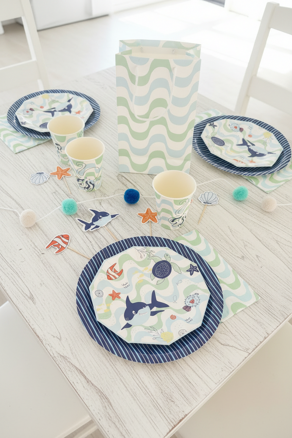 A coordinated ocean-themed party table setting with printed paper plates, cups, napkins, starfish cutouts, and pom-pom garlands on a wooden table.